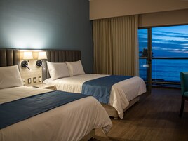 Habitación de hotel con dos camas, un ventanal con vista al océano y un balcón con una silla.