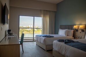 Habitación de hotel con dos camas, un escritorio con una computadora portátil y cafetera, una silla y un ventanal con vista a una masa de agua y edificios.