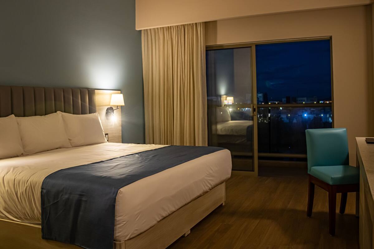 A hotel room with a large bed, a desk with a chair, a window with a view of the city, and a curtain.