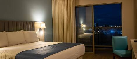 A hotel room with a large bed, a desk with a chair, a window with a view of the city, and a curtain.