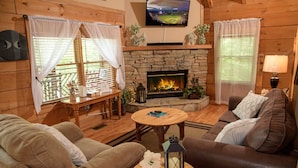 A cozy living room with a stone fireplace, comfortable sofas, a television mounted on the wall, and a view of a baseball field through the windows. - mountain view log cabin in Gatlinburg (Gatlinburg)