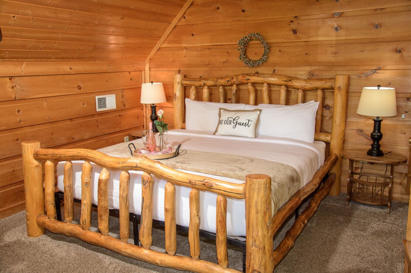 A wooden cabin bedroom with a bed, two bedside lamps, a small table with a vase, and a 'BE OUR GUEST' pillow.