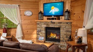 A cozy cabin interior with a stone fireplace, leather sofa, and a television displaying the weather. - Cuddley Bears hideaway (Sevierville)