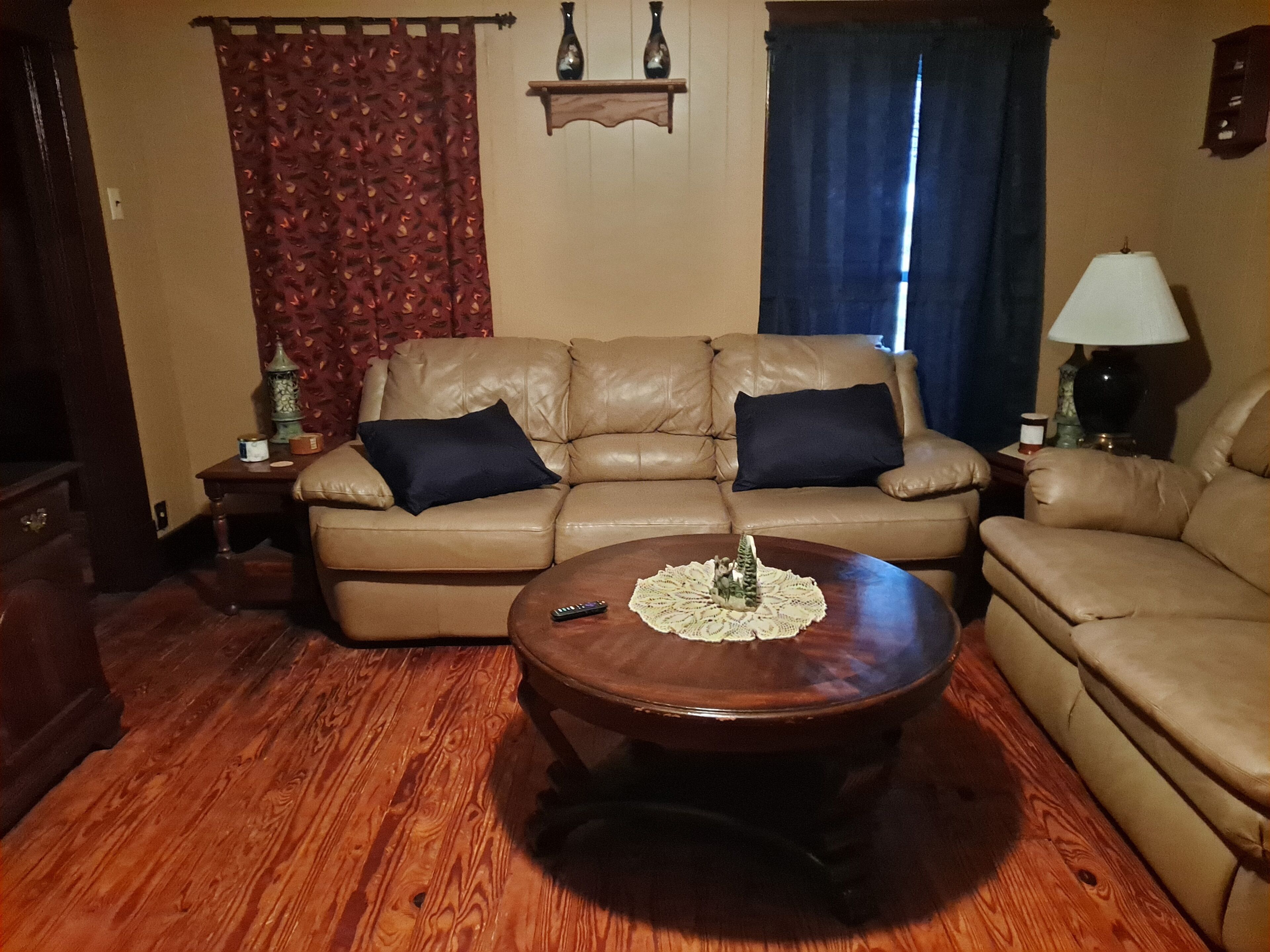 A living room with a brown leather sofa, two black cushions, a wooden coffee table with a lace doily, a remote control, and a small Christmas tree.