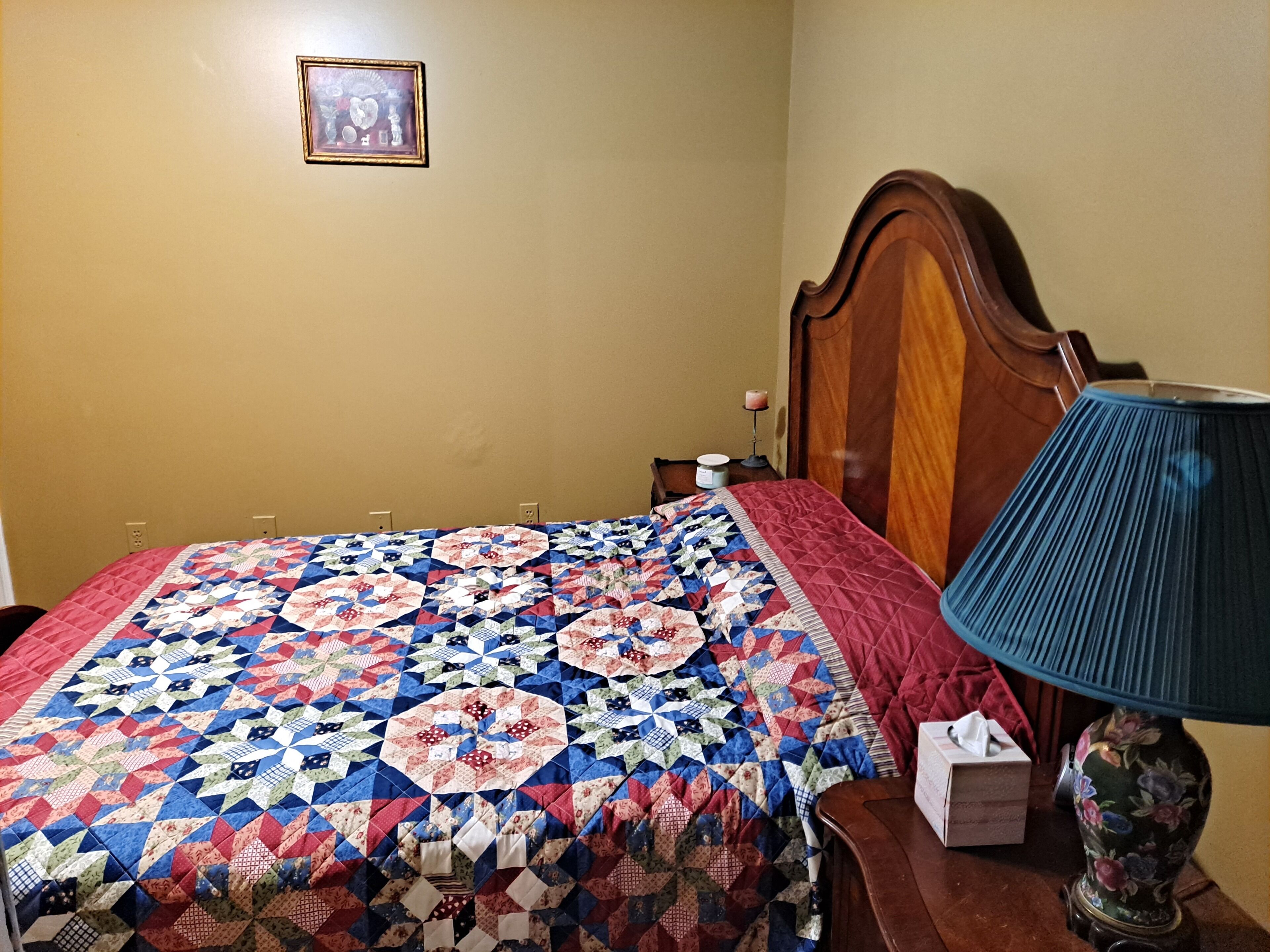 A bedroom with a wooden headboard, a quilted bedspread, a bedside table with a lamp, and a framed picture on the wall.