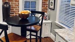 Un coin repas avec un mur en briques, une table ronde entourée de chaises, une horloge et une fenêtre avec des stores.