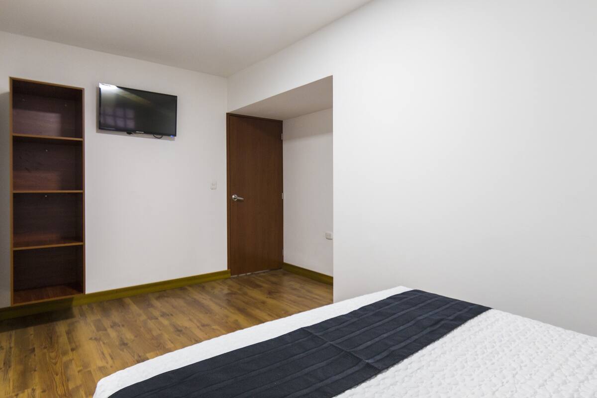 A bedroom with a wooden floor, a bed with a white and black bedspread, a television mounted on the wall, and a wooden wardrobe.