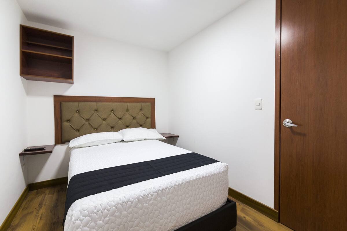 A bedroom with a bed, wooden headboard, bedside tables, a wardrobe, and a shelf.