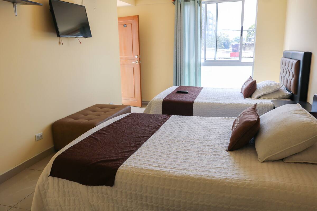 A hotel room with two beds, a television on the wall, and a window with curtains.