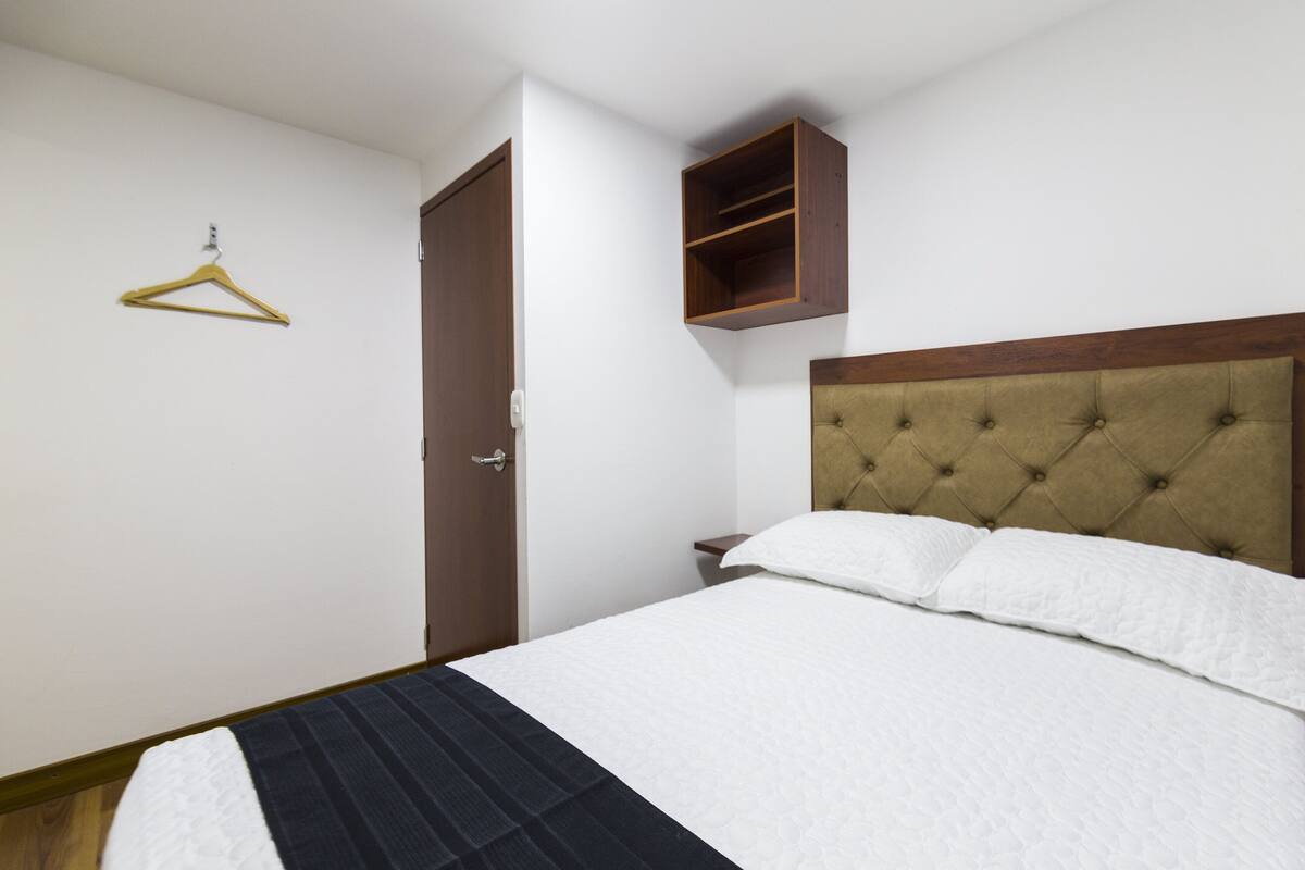 A neatly made bed with a tufted headboard, a wooden bedside table, and a hanging garment rack.