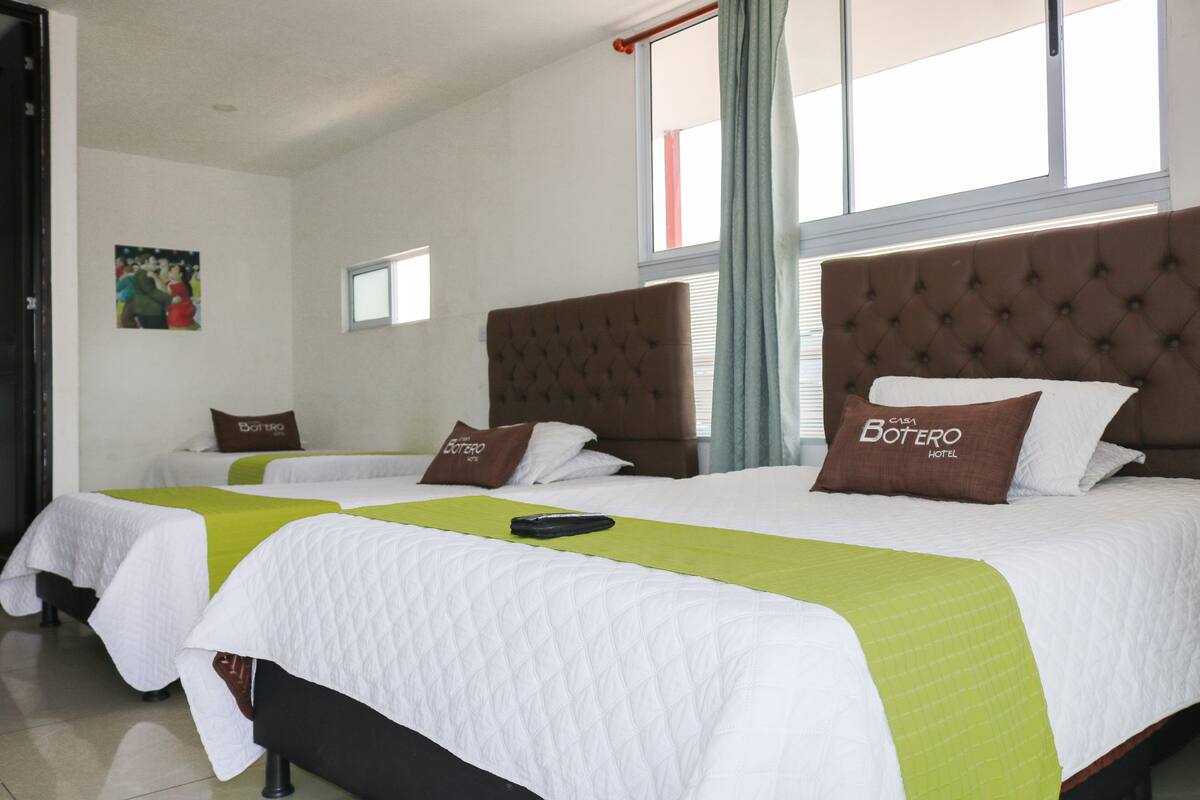 A hotel room with three beds, each with a green and white bedspread and a brown headboard. There is a painting on the wall and a window with curtains.