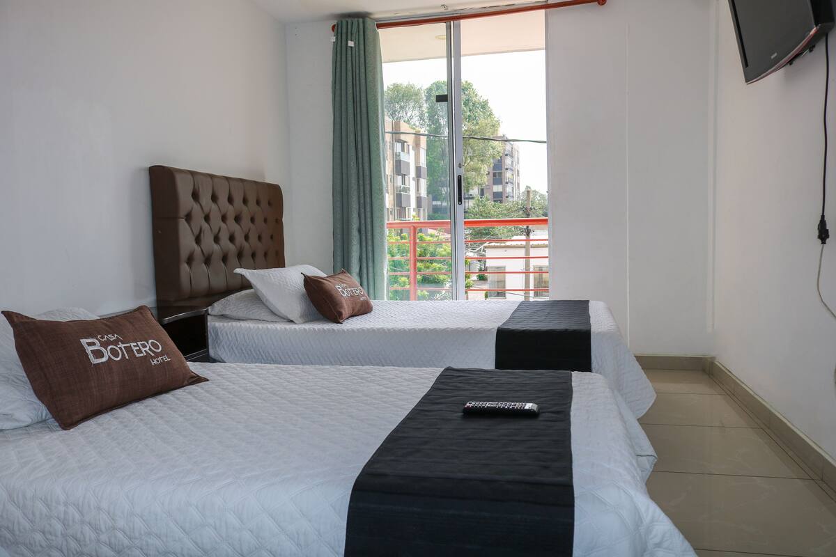 A hotel room with two beds, a TV mounted on the wall, and a view of the outside balcony.