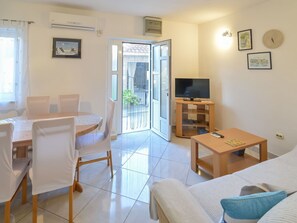 Un salon comprenant un canapé, un téléviseur posé sur un meuble, une table à manger avec des chaises et une porte-fenêtre coulissante donnant sur un balcon.