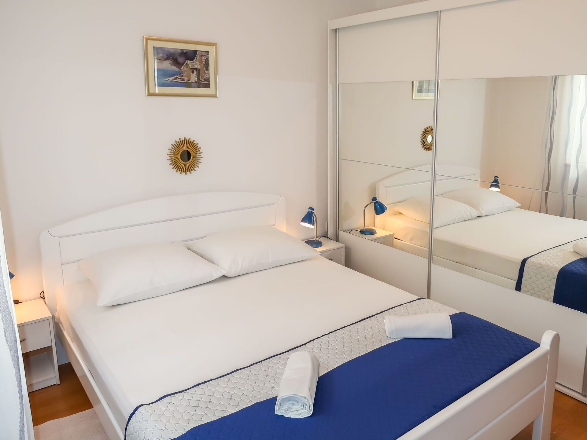 A neatly arranged bedroom with a large mirror, a bedside lamp, and a painting on the wall.