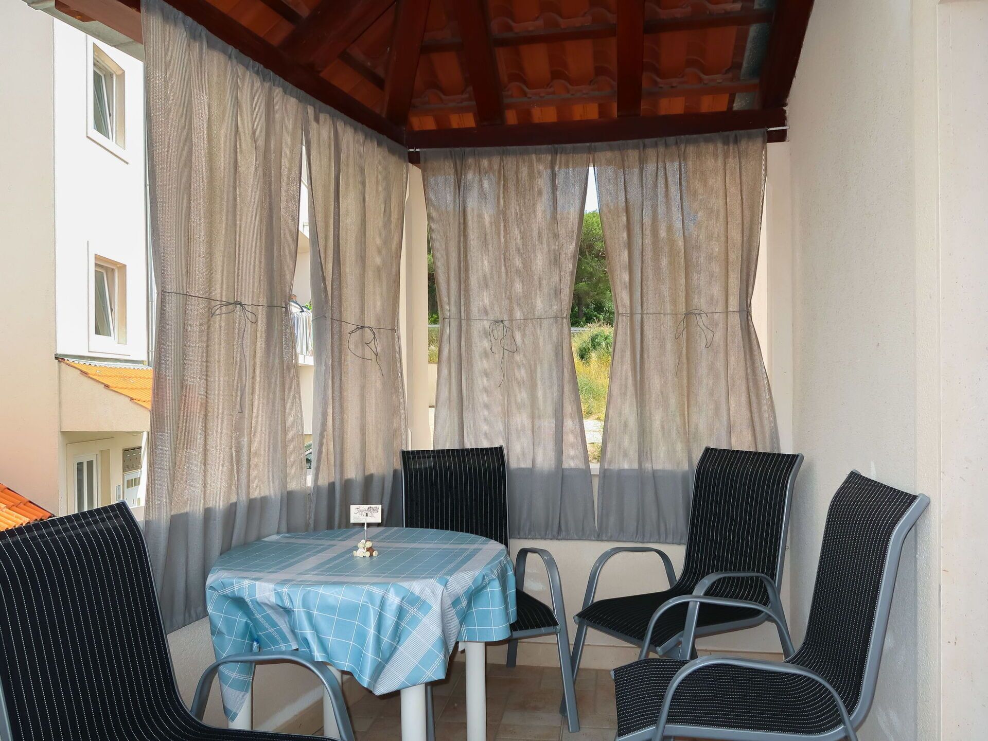 A covered outdoor seating area with a table and four chairs, a view of greenery through the windows, and a building in the background.