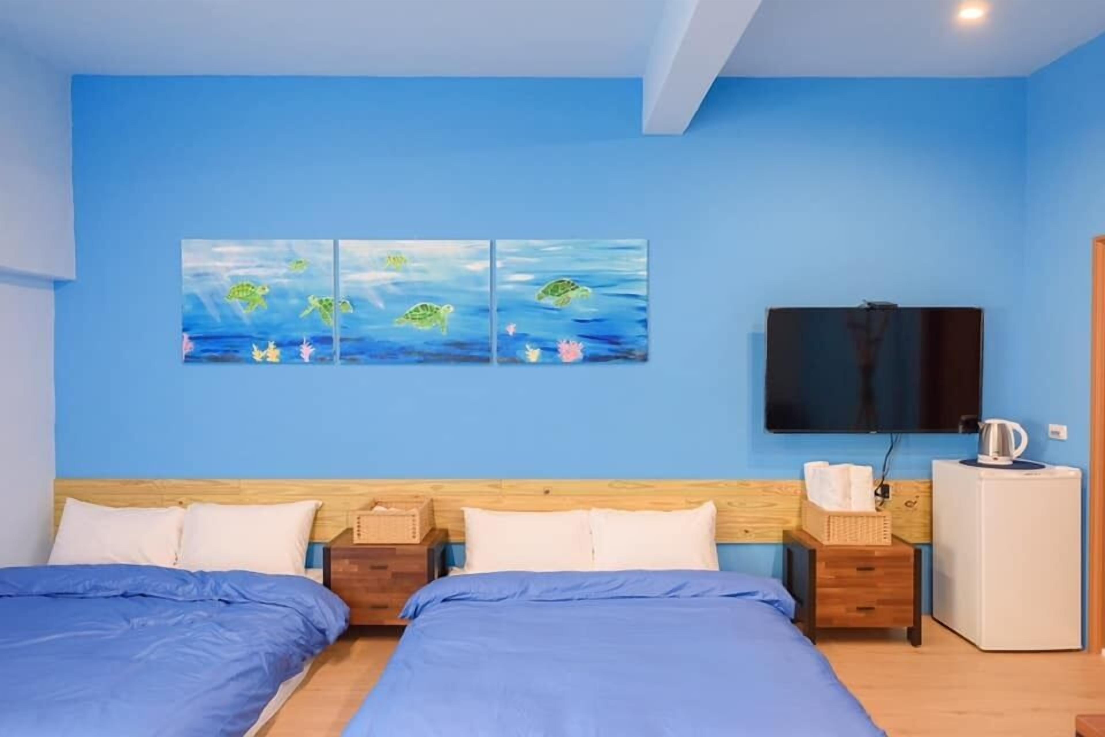 Quarto com duas camas, televisão de tela plana, pequeno frigorífico e obras de arte na parede.