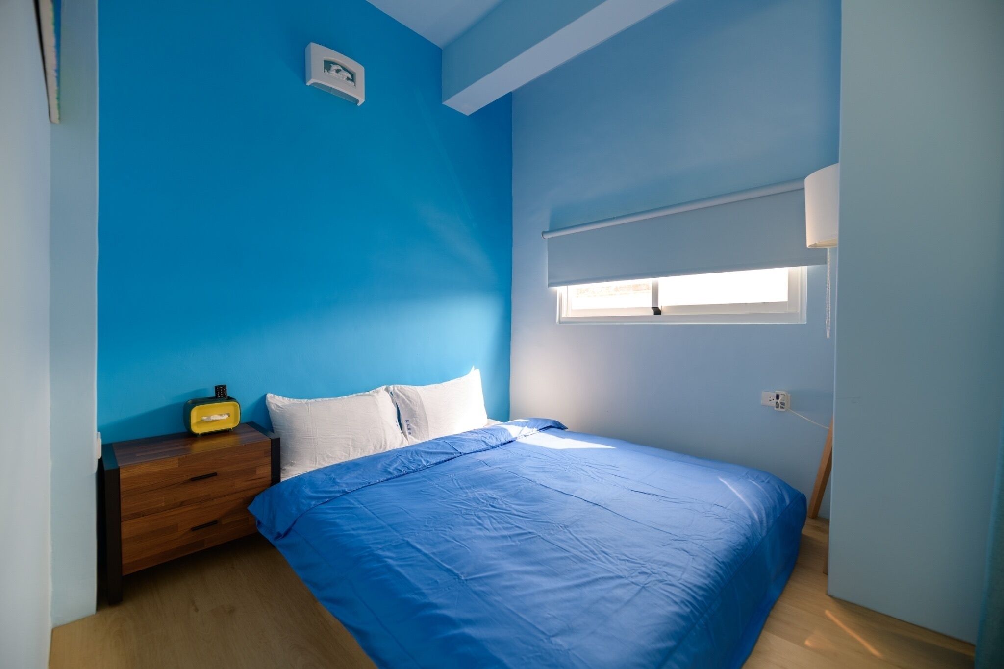 A bedroom with a blue wall, a wooden nightstand with a radio, a bed with blue bedding, and a window with blinds.