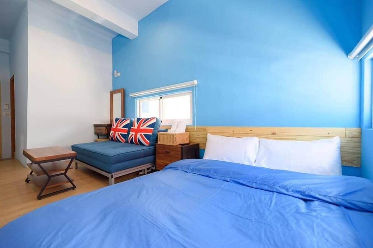 Une chambre à coucher avec un lit, une table de chevet, une commode, une fenêtre avec des stores et un mur bleu.