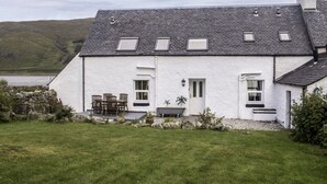 Une maison blanche à deux étages, avec un toit en pente, un petit porche doté d’une table et de chaises, et une vue sur un lac.