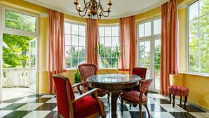A dining area with a round table and chairs, large windows with curtains, and a chandelier.