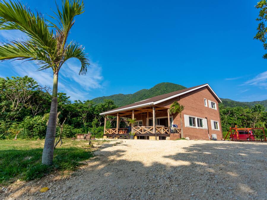 A two-story brick house with a covered porch, surrounded by greenery and hills.