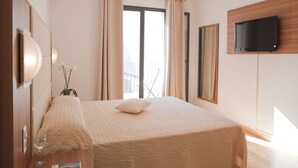 Habitación de hotel con una cama, un televisor montado en la pared, una ventana con cortinas y una mesita de noche con un vaso de agua y un libro.