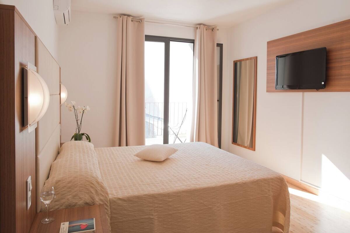 A hotel room with a bed, a TV mounted on the wall, a window with curtains, and a bedside table with a glass of water and a book.