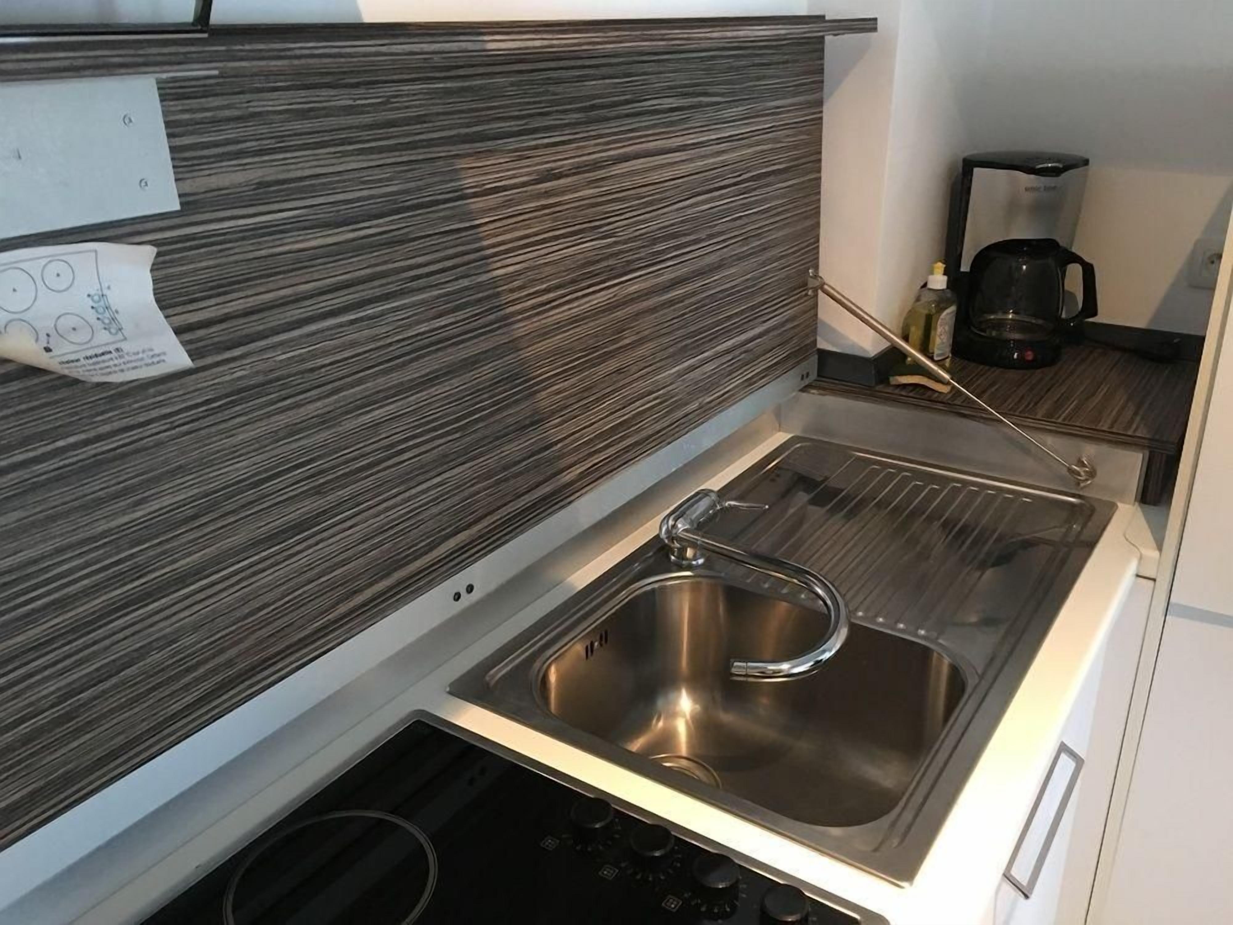A compact kitchen with a stainless steel sink, a coffee maker, and a paper with a drawing on the wall.