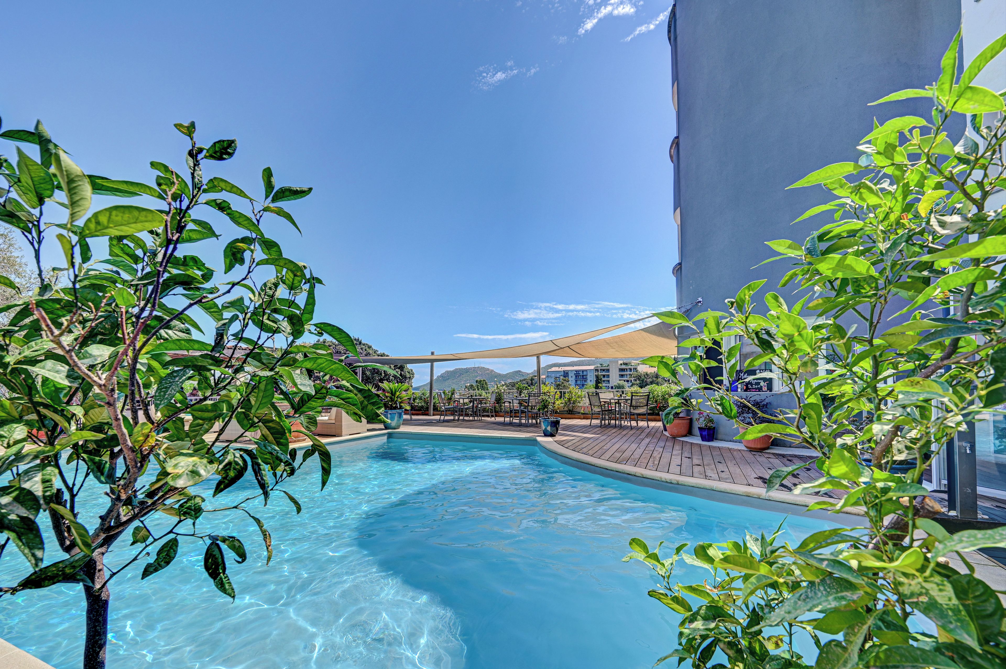 A clear blue pool surrounded by greenery and a wooden deck with outdoor seating.