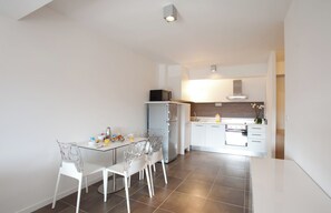 Une cuisine moderne avec des meubles blancs, un réfrigérateur et un coin repas comprenant une table et des chaises en verre.