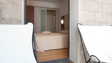 A room with a bed, two white chairs, and a view into another room with a bed and a nightstand.