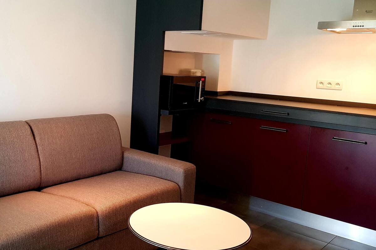 A modern living room with a sofa and a small round table, a kitchen area with dark cabinets and a microwave, and a white wall with a light switch.