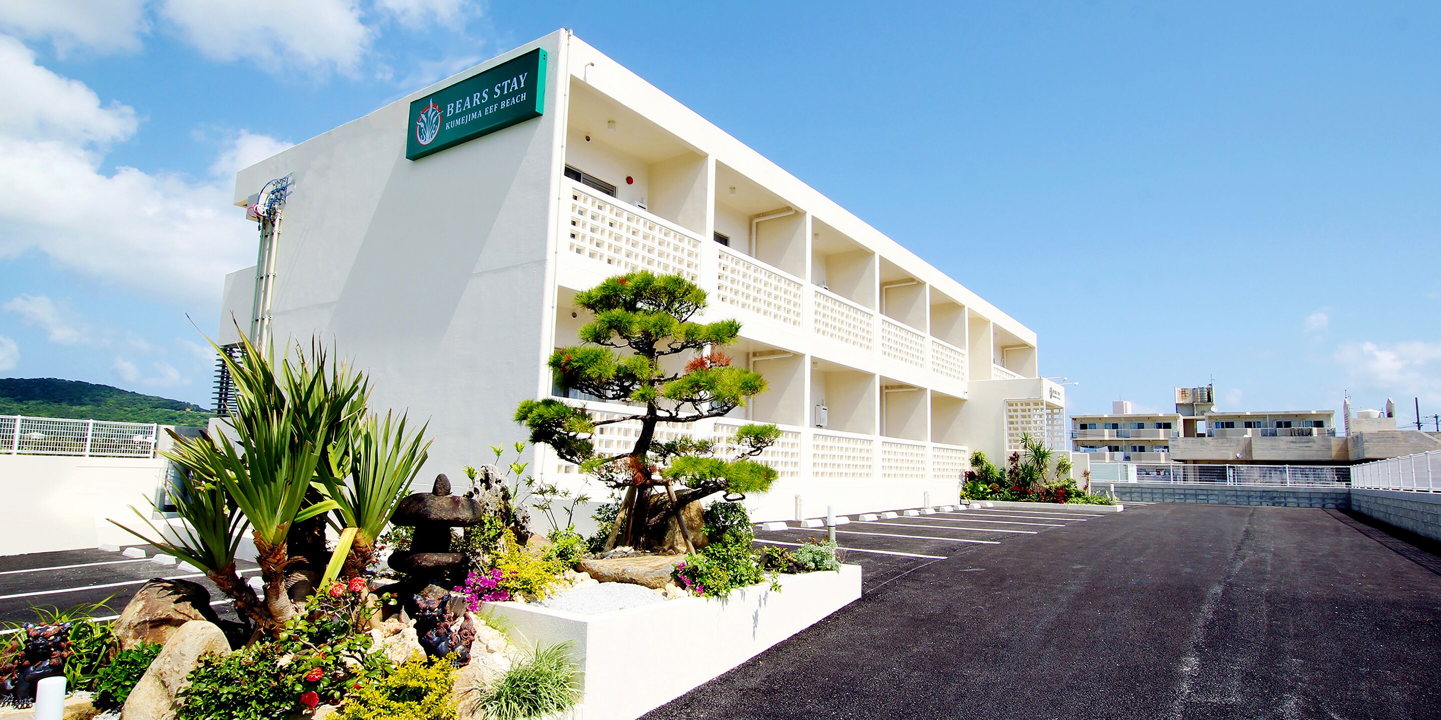 Un hotel de varios pisos con fachada blanca, un cartel que dice "BEARS STAY KUMAMURA BEP BEACH" y un estacionamiento frente.