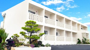 Un hôtel à plusieurs étages, à la façade blanche, doté de balcons et d’un jardin bien entretenu où l’on trouve diverses plantes et un bonsaï.