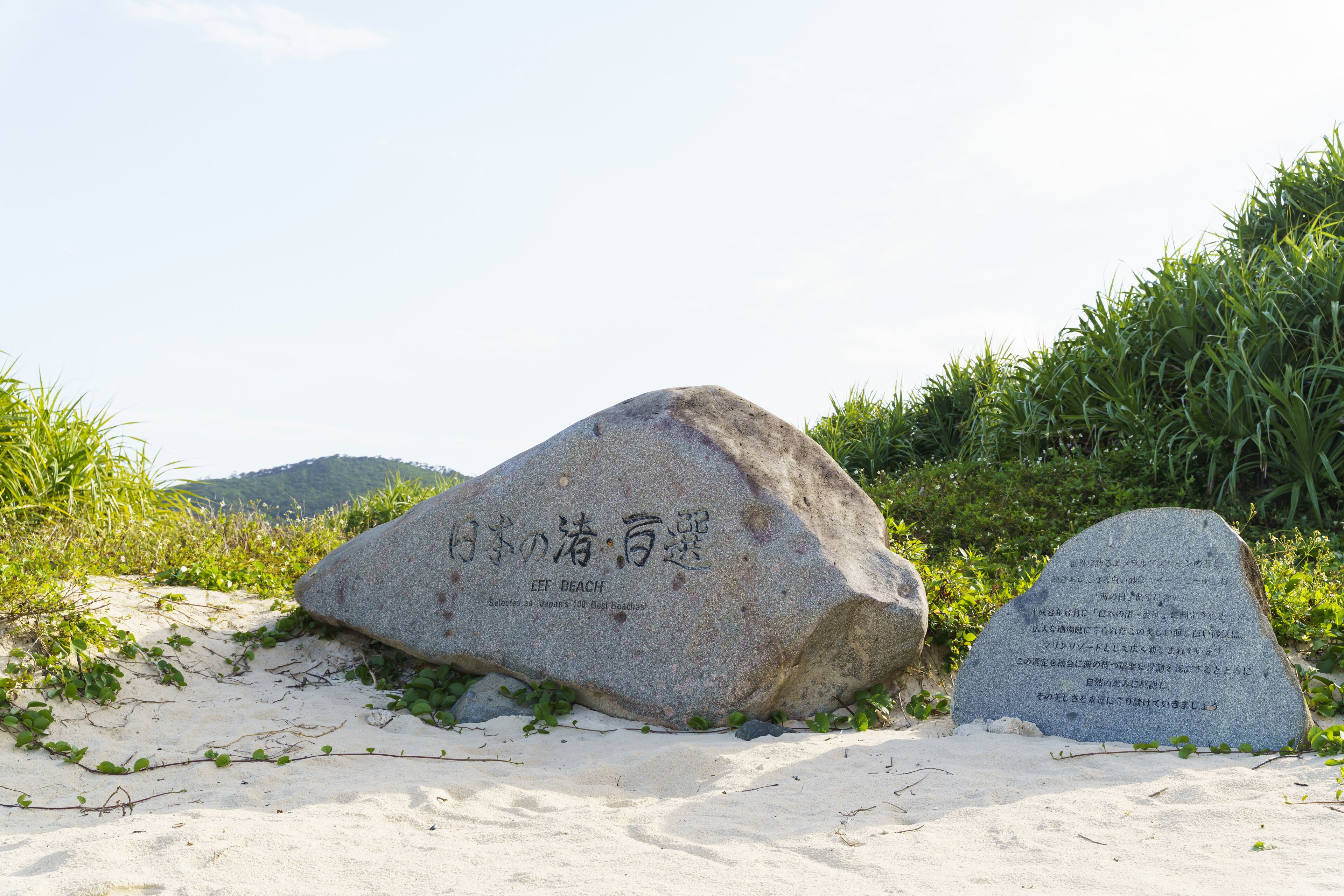 A large rock with Japanese text and a smaller stone with more text, both on a sandy beach with greenery in the background.