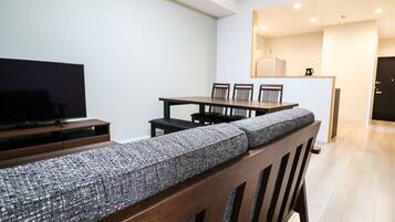 A modern living room with a sofa, a TV, a dining table with chairs, and a kitchen area in the background.