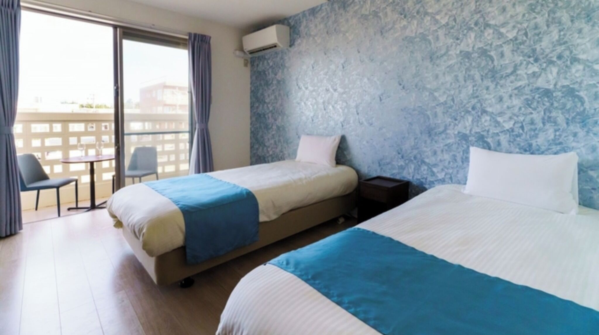 Ein Hotelzimmer mit zwei Betten, einem kleinen Tisch, einem Stuhl und einem Balkon mit Blick auf Gebäude.