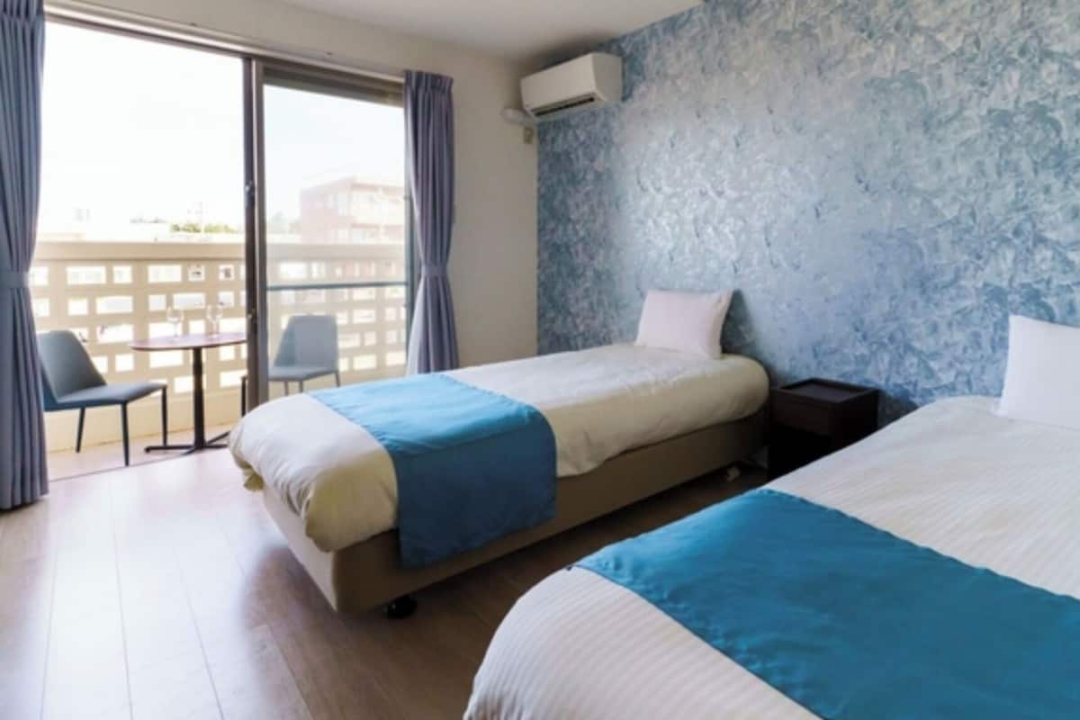 Une chambre d’hôtel avec deux lits, une petite table, une chaise et un balcon donnant sur des immeubles.