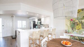 A kitchen with white cabinetry, a dining area with a round wooden table, and a large window. - The Cocohut Sanibel at Loggerhead Cay 🐚 Beachfront Condo with Pool & Pickleball (Sanibel)