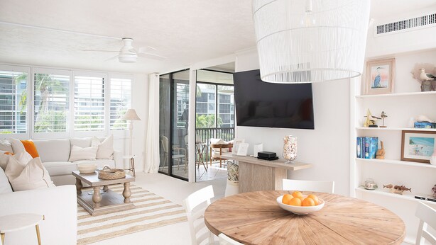 A bright living room with a white sofa, a wooden dining table with chairs, a TV mounted on the wall, and a balcony with a view of greenery. - The Cocohut Sanibel at Loggerhead Cay 🐚 Beachfront Condo with Pool & Pickleball (Sanibel)