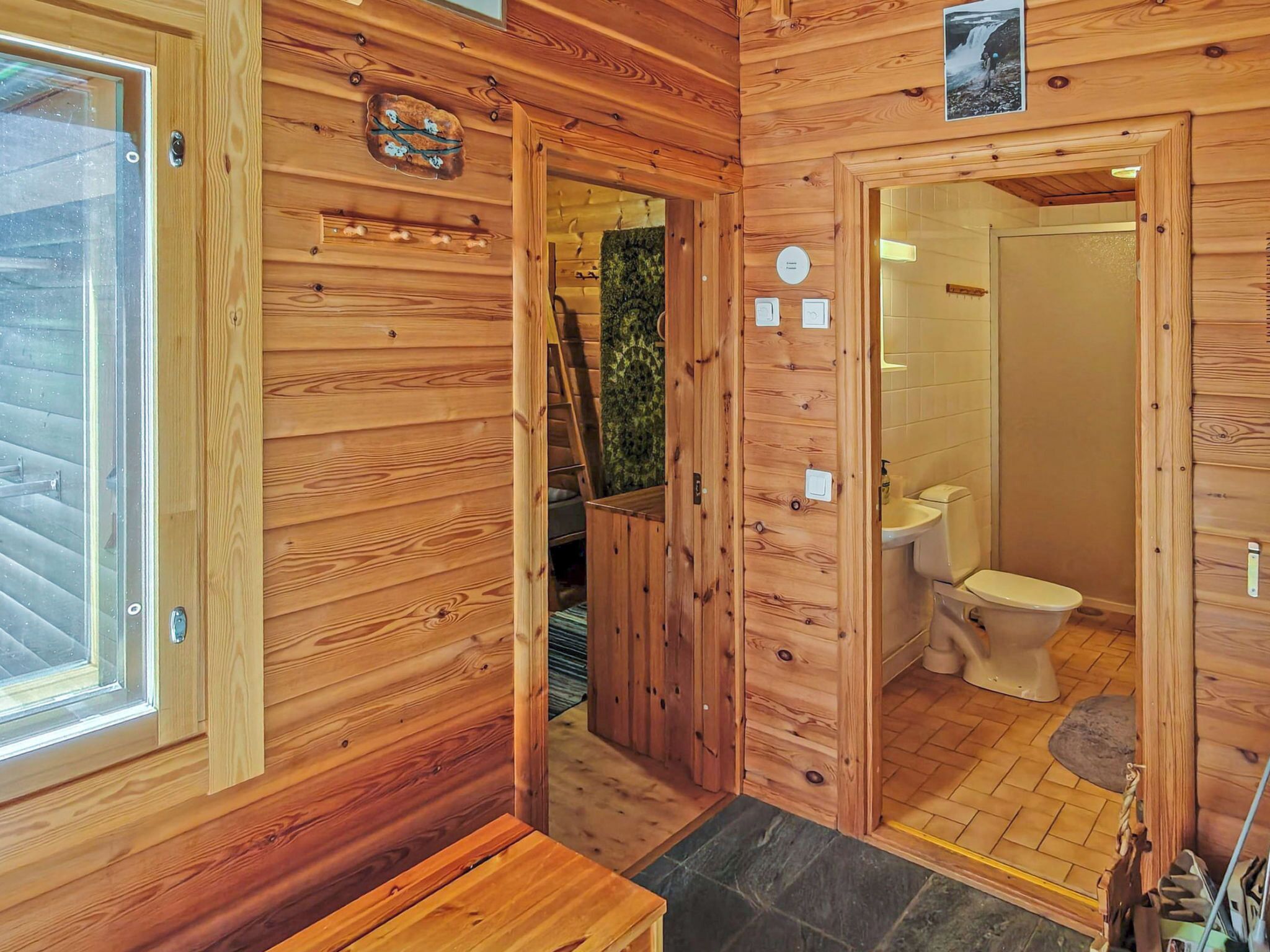 Interior de una cabaña de madera con puerta de madera, una ventana, un baño con inodoro y un pequeño banco de madera.