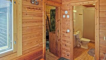 Interior de una cabaña de madera con puerta de madera, una ventana, un baño con inodoro y un pequeño banco de madera.