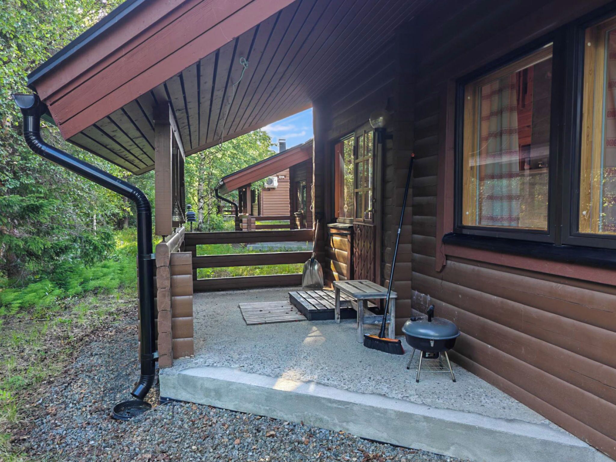 Une cabane en bois dotée d’un porche couvert, d’un barbecue et d’une fenêtre donnant sur une zone boisée.