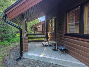 A wooden cabin with a covered porch, a barbecue grill, and a window overlooking a forested area.