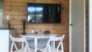 A dining area with a white table and chairs, a flat-screen TV mounted on the wall, and a wooden ceiling. - Barbs Bach (Mangawhai)