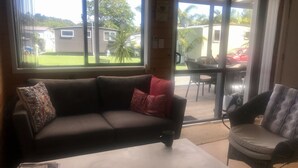 A living room with a sofa, a coffee table, and a chair. There is a window with a view of a house and a car. - Barbs Bach (Mangawhai)