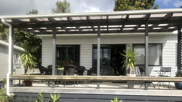 A covered outdoor seating area with a pergola, potted plants, and a stone border. - Barbs Bach (Mangawhai)