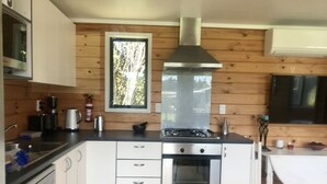 A compact kitchen with a stove, oven, and sink, wooden paneling, and a window with a view of trees. - Barbs Bach (Mangawhai)
