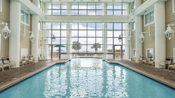An indoor swimming pool with clear glass walls, lounge chairs, and a view of palm trees outside.