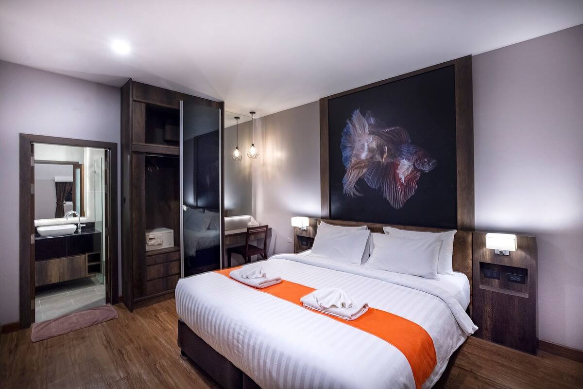 Habitación de hotel con cama, un espejo grande, escritorio, silla y mesita de noche con lámpara.